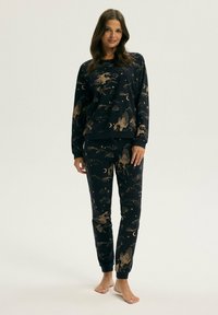 Kobieta stoi boso, ubrana w pasujące czarne długie rękawy i spodnie z złotym wzorem pegaza, księżyca i chmur w piżamie.