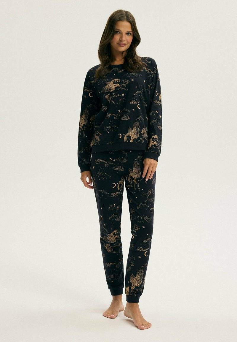 Kobieta stoi boso, ubrana w pasujące czarne długie rękawy i spodnie z złotym wzorem pegaza, księżyca i chmur w piżamie.