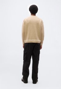 Άνδρας με κολεγιακό κούρεμα αφρο, φοράει μπεζ πουλόβερ και μαύρο παντελόνι cargo, στέκεται με την πλάτη προς τον θεατή μπροστά σε απλό λευκό φόντο.