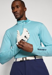 Jasnoniebieska, długorękawowa koszulka sportowa z wysokim kołnierzem, w połączeniu z białymi rękawicami golfowymi z czarnymi akcentami. Granatowy pasek z metalową klamrą.