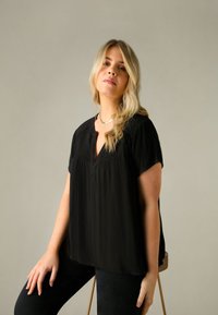 Svart kortärmad topp med texturerad tyg och en subtil V-ringning, tillsammans med mörka jeans, placerad mot en neutral bakgrund.