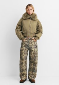Skrajšana olivno zelena bomber jakna z umetnim krznom na ovratniku, kombinirana s širokimi hlačami z leopardjim vzorcem v bež in rjavih tonih.