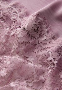Spitzenstoff in sanftem Lavendel mit filigranen Blumenmustern und zarten, geschwungenen Kanten auf einer glatten, leicht glänzenden Basis.
