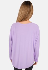 Lavendelfarbenes Langarmshirt mit rundem Ausschnitt und lässiger Passform. Weiches Material, leicht drapierter Rücken und gewellter Saum. Schwarze Hosen sind sichtbar.