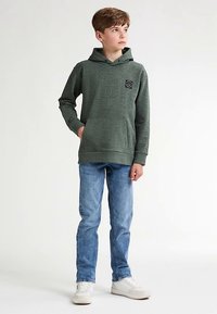 Zelená mikina s kapucí s reliéfním motivem, černá štítek s logem, doplněná světle modrými džínami a bílými teniskami. Hladká textura, volný střih.