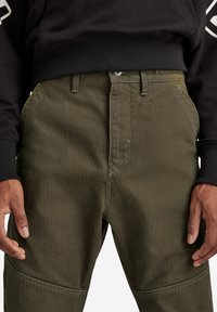 Olivgröna byxor med texturerat tyg, synliga sömmar och rak skärning. Har bälteshällor och framfickor. Svart sweatshirt ovanför.