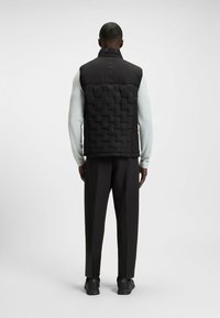 Svart ärmlös puffer-väst med en texturerad quiltad design, ståkrage och slät tyg på den övre delen, kombinerad med svarta byxor.