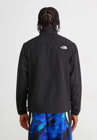Person med fletninger iført en sort The North Face jakke og blå mønstrede shorts, vender væk mod en ensfarvet baggrund.