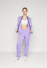 Šviesiai violetinis sportinis kostiumas su baltais akcentais, sudarytas iš zip-up striukės ir elastingų kelnių, derinamas su baltu sportiniu liemenėliu ir bateliais.
