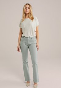 Gai bēšas īsām piedurknēm blūze ar horizontālu tekstūru, saskaņota ar gaiši zaļām platas kājas biksēm un bēšu blokpēdu kurpēm. Neitrāls fons.