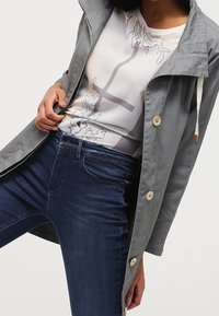 Šedá lehká bunda s kapucí na stažení, knoflíky a hladkým povrchem, nošená přes vzorovanou bílou košili a tmavě modré džíny.