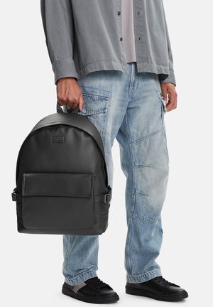 Uomo tiene uno zaino in pelle nera per la maniglia superiore, indossa jeans cargo blu chiaro, maglietta grigia e sneakers nere su sfondo bianco.