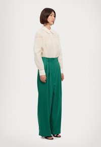 Blusa cream con pieghe verticali e bordo in pizzo, abbinata a pantaloni verde ampia a vita alta. Indossati con sandali neri.