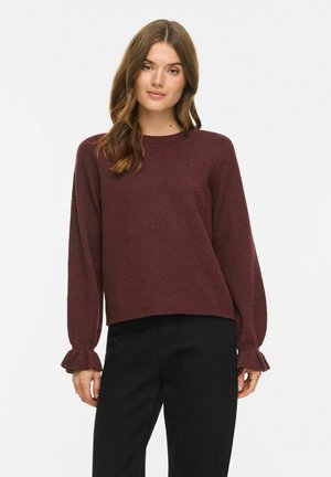 Femme aux cheveux bruns ondulés portant un pull bordeaux à manches longues avec des poignets volantés et un pantalon noir, debout sur un fond blanc uni.