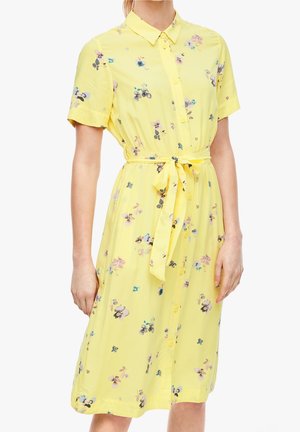 Vestito giallo a maniche corte con colletto, chiusura con bottoni sul davanti, motivo floreale e cintura annodata in vita, indossato da una donna in piedi mostrata dal collo alle ginocchia.
