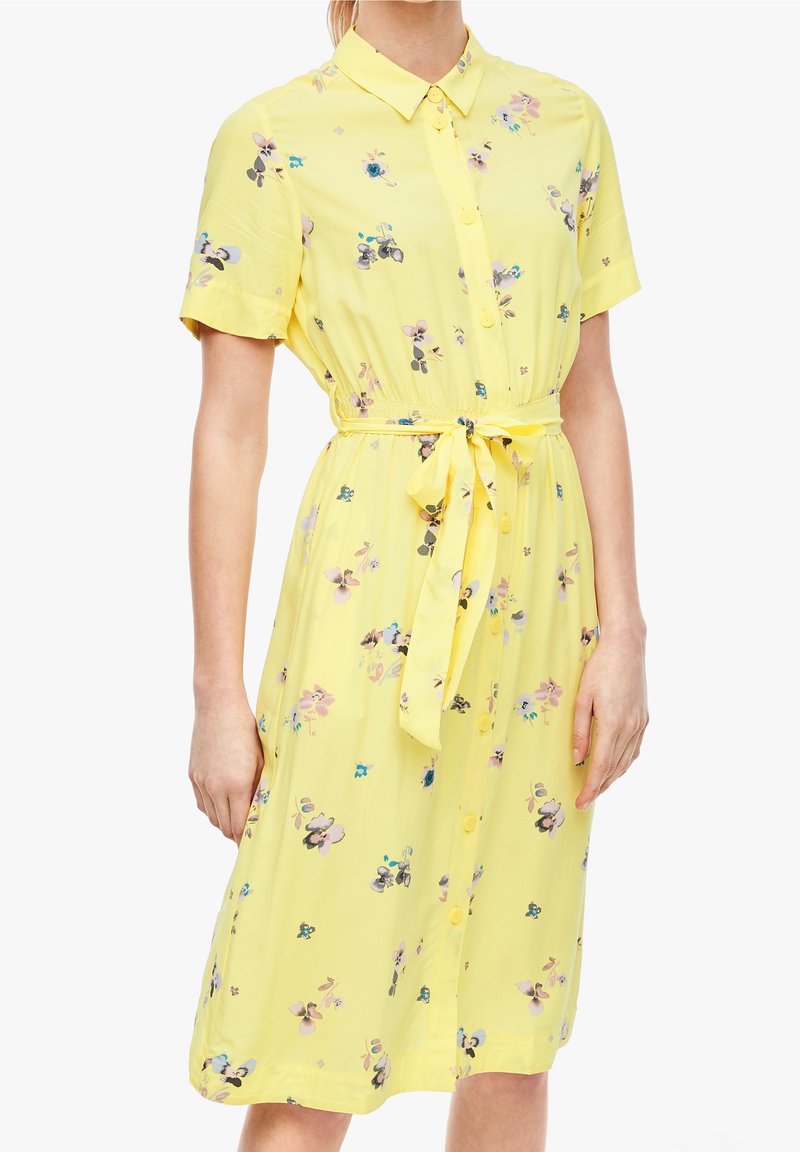 Robe jaune à manches courtes avec col, boutonnée sur le devant, motif floral et ceinture nouée à la taille, portée par une femme debout visible du cou jusqu'aux genoux.