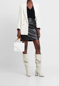 Žena v bílém blejzru, černém topu, černé kožené sukni, bílých botách a s bílou kabelkou s řetízkem z želvoviny.