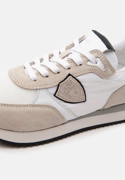 Basket blanche et beige avec des empiècements en suède, des lacets blancs, un logo à bordure noire sur le côté et une semelle texturée, visible sur fond blanc.