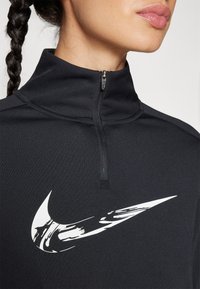Black zippered top with a high collar, made of smooth fabric. Features a prominent white Nike swoosh logo on the chest.