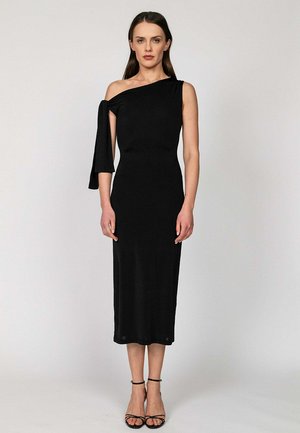 Kobieta w czarnej midi sukience na jedno ramię z drapowanym rękawem na lewej ręce, stojąca na jednolitym jasnym tle.