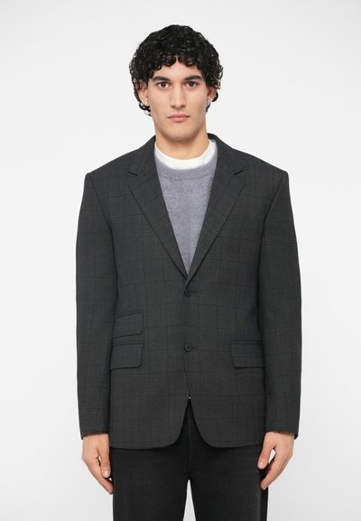 Blazer gris foncé à carreaux avec deux poches avant, revers crantés et fermeture par un seul bouton, porté sur un pull gris clair.
