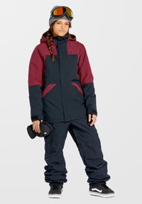 Snowboardjakke med maroon og sorte farveblokke, justerbar hætte og lynlåslommer. Matchet med løse sorte skibukser og sorte sneakers.