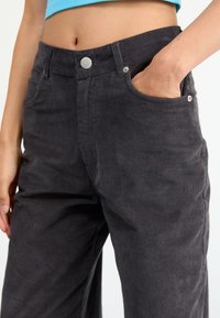 Gros plan sur une personne portant un pantalon en corduroy noir taille haute, avec une main dans la poche avant et un ventre visible au-dessus de la taille.