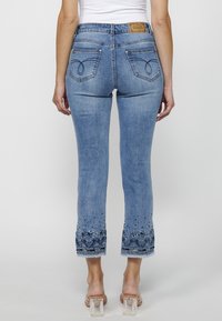 Jean en denim bleu clair avec une coupe ajustée, ourlet orné d'un motif floral et étiquette sur la ceinture. Cinq poches visibles.