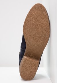 Botín de ante azul marino con suela de goma texturada, detalle de cremallera lateral y un tacón redondeado ligeramente elevado. Talla marcada como 37.