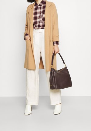 Braune Lederhandtasche mit einem lässigen Design, goldfarbene Hardware-Akzente, vor einem hellen Outfit, das mit einem beige Mantel und einem karierten Hemd kombiniert ist.