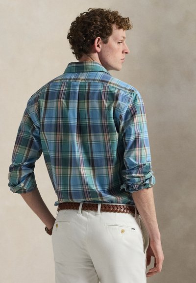 Checkered shirt in blue, green, and pink hues; button-down collar; rolled sleeves; complemented by a braided brown belt and white pants.