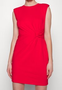 Röd ärmlös klänning i mjukt tyg, med en tvistad knutdetalj vid midjan och en rund halsringning. Fitted silhuett.