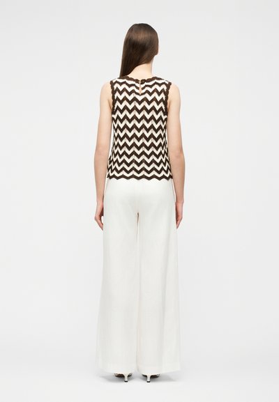 Γυναίκα με μακριά ίσια μαλλιά που φοράει αμάνικο μπλουζάκι σε ασπρόμαυρο σχέδιο ζιγκ-ζαγκ και λευκό παντελόνι με φαρδιά μπατζάκια, στέκεται γυρισμένη πλάτη.