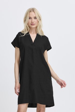 Femme blonde portant une robe noire ample à manches courtes, debout devant un fond blanc uni.