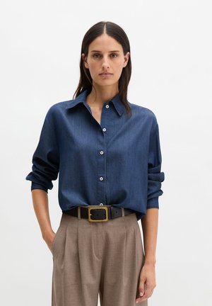 Femme aux cheveux noirs raides portant une chemise bleue à boutons avec les manches retroussées et un pantalon beige taille haute avec une ceinture noire.