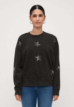 Kvinne med mørkt hår i hoopsmykker har på seg en svart hettegenser dekorert med hvite stjerne mønstre og blå jeans, stående mot en ensfarget bakgrunn.