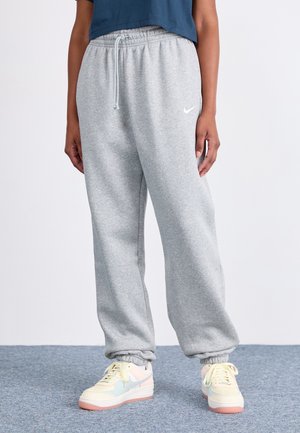 Osoba w luźnych szarych spodniach dresowych Nike z elastycznymi mankietami i wielokolorowych sneakersach Nike stojąca na szarym wykładzinowym dywanie.