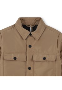 Tan quilted jacket with a collared neckline, two chest pockets with black buttons, and a smooth, shiny texture.