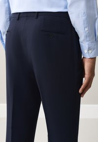 Uomo che indossa pantaloni eleganti blu navy e una camicia azzurro chiaro a maniche lunghe, vista posteriore dalla vita a metà coscia.