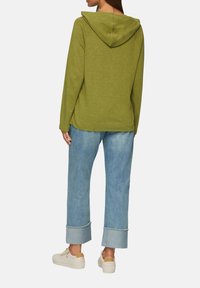 Olivgrüner Hoodie mit Kapuze am Nacken, kombiniert mit hellblauen, cropped Jeans mit fransigem Saum und weißen Turnschuhen.