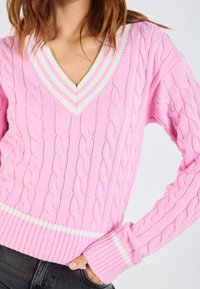 Roze kabelgebreide trui met V-hals, voorzien van witte gestreepte accenten op de kraag en zoom. Geribde boorden en zoom. Gelaagd patroon door de hele trui.