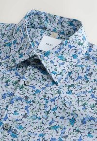 Camisa con estampado floral hecha de tela ligera, con base azul claro y detalles en blanco, verde y azul. Cuello abotonado y dos botones.