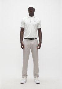 Άνδρας όρθιος, φοράει λευκό πόλο, ανοιχτό γκρι παντελόνι, λευκά αθλητικά παπούτσια, μαύρη ζώνη και λευκό καπέλο, μπροστά σε απλό λευκό φόντο.