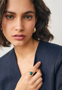Goldring mit einem grünen Stein, am Finger getragen. Modell in einem dunkelblauen Kleid, mit natürlichem Makeup und lockigem Haar. Neutraler Hintergrund.