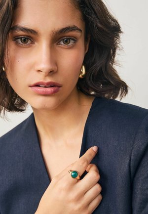 Goldring mit einem grünen Stein, am Finger getragen. Modell in einem dunkelblauen Kleid, mit natürlichem Makeup und lockigem Haar. Neutraler Hintergrund.