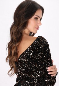 Czarna sukienka z błyszczącymi detalami z cekinów, prezentująca fason na ramiączkach i fakturowany materiał, uzupełniona długimi kolczykami.
