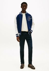 Chaqueta varsity azul y blanca con el logo "F" bordado, combinada con un suéter crema, jeans oscuros y mocasines marrones.