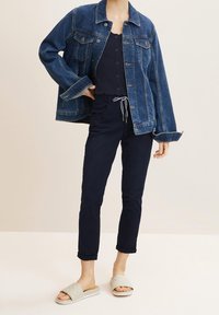 Niebieska kurtka dżinsowa o luźnym kroju, zestawiona z ciemnym topem na ramiączkach, granatowymi spodniami podwiniętymi u dołu oraz jasnoszarymi pikowanymi klapkami.