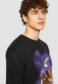 Svart bomull T-skjorte med et fargerikt trykk av en basketballspiller på forsiden, med abstrakte elementer i lilla og gult. Rund hals design.