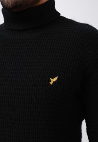 Suéter negro de punto con cuello alto, con un patrón texturizado y un pequeño logotipo de un pájaro dorado en el área del pecho izquierdo.